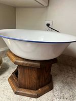 White enamel metal dish bowl with blue rim, shown from a side angle on a counter with wooden stand beneath, showing profile and size.