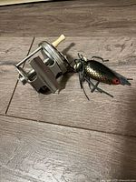 Fishing reel and fishing lure side by side on wood floor showing metal reel with ivory handles and black lure with rubber skirts and red eyes.