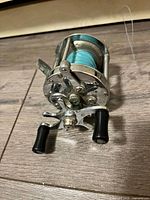Fishing reel showing side with black crank handles and blue fishing line wound on spool.