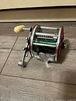 Side view of vintage fishing reel showing crank handle, spool wound with green fishing line, and red side plate.