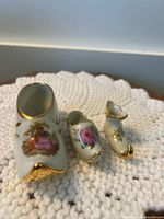 Front view of three porcelain shoes with floral and angel designs, gold accents