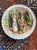 Front view of the ceramic musical plate showing detailed 3D scene of Peter Rabbit and Benjamin Bunny in garden with vegetables and greenery, white and blue border.