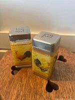 Pair of vintage glass salt and pepper shakers with rooster illustrations on yellow painted background and metal tops.