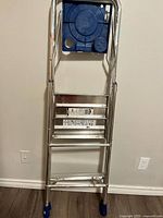 Step ladder folded standing against a wall, showing front side with four aluminum steps and blue top tray.