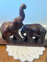 Front view of two wood carved elephants on a base, one with trunk raised, the other with trunk down. Both missing tusks except one.