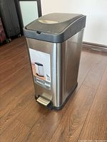 Rectangular stainless steel step trash can with black lid and base, foot pedal in front, placed on wooden floor.
