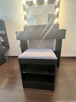 Photo showing black and gray wooden dressing table with lighted mirror, cushioned stool, and open storage shelves below stool.