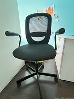 Black computer chair with mesh backrest and fabric seat shown front-facing in a room between a white wall and white desk.