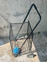 Black metal utility cart with wire mesh basket, two wheels, and ergonomic handle placed against a concrete wall.