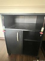 Black wood composite storage cabinet showing one closed door with silver handle, an open shelf above the door, and open compartments to the side.