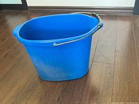Blue plastic cleaning bucket with sturdy plastic handle sitting on wooden floor, showing minor scratches and wear.