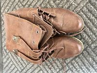 Top view of a pair of brown leather knee-high lace-up boots placed on carpet showing details of the leather texture, eyelets, and laces.
