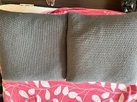 Photo shows two square grey cushions with a knit textured fabric, placed side by side on a pink and white leaf patterned surface.