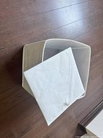 Medium-sized cream fabric collapsible storage box shown open and placed on dark wood floor, displaying its foldable structure and rectangular shape.