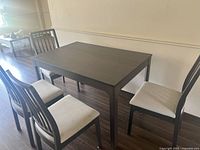 Photo of a dark brown wood rectangular dining table with four matching chairs with light beige fabric upholstered seats, showing the full set against a neutral wall and hardwood floor.