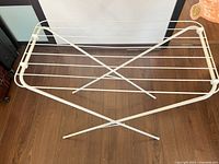 Top view of the white foldable metal clothes drying rack open and standing on wooden floor, showing the multiple horizontal bars and X-frame support.