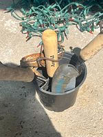 Top-down photo showing garden tools with wooden handles and rusted metal parts inside a black plastic bucket on concrete ground. Green Christmas lights in the background.