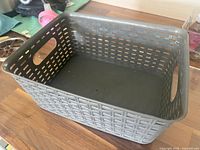 Photo of a gray plastic rectangular storage basket with ventilated sides and handles, placed on a wooden surface with minor background items.