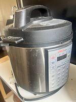 Front angled photo showing Instant Pot style electric pressure cooker with digital control panel, handle, and safety lock lid with visible dust accumulation.