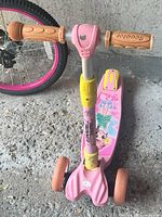 Front view of a pink kids' scooter showing the two wheels, beige textured handles, yellow and pink column, cartoon character decal on the foot deck, and textured foot brake.