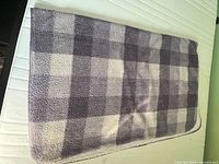 Photo showing a large fleece blanket folded over a white mattress or surface. The blanket has a blue and white plaid pattern with various shades of blue forming the checkered design and appears soft and thick.