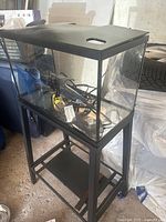 Medium-sized rectangular glass aquarium on black metal stand with lower shelf, black lid with openings, shown with aquarium accessories inside including filter, heater, cables, and lighting unit.