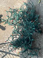 Photo showing a tangled strand of multi-color Christmas lights with green wiring. Multiple small incandescent bulbs in red, orange, green colors are visible. The strand appears moderately long and is suitable for indoor or outdoor use as per seller.