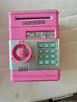 Front view of pink plastic piggy bank shaped like a safe, showing keypad with numbers and lock dial, coin and bill slots on top.