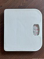 Top-down view of white mechanical bathroom scale on hardwood floor, showing ridged surface and analog dial.