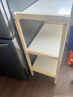 Single photo showing the storage rack placed beside a refrigerator with three white shelves and light wood frame structure.