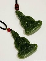 Close-up photo of one green jade Buddha pendant lying flat on a white surface showing detailed carving and attached cord with wooden bead.