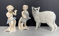 Two Goebel cherub figurines and one white ceramic cat figurine on black background.