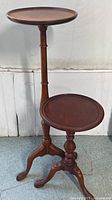 Two vintage mahogany plant stands of varying heights next to wall, showing round tops and turned pedestal bases with claw feet.