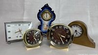 Group photo showing five clocks: two Westclox Big Ben alarm clocks, one rectangular white clock with silver trim, one wooden mantel clock, and one blue and white porcelain mantel clock with cherub decoration.