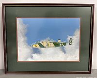 Full view of framed aircraft artwork showing the plane in flight against a blue sky with clouds, mounted in an olive green mat and dark wood frame.