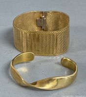 Two gold tone bracelets displayed together: a thick woven mesh bangle and an open cuff with a twisted design on a neutral surface.