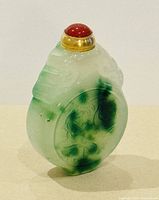 Front view of carved jade snuff bottle with brass and red stone stopper. Shows translucent jade with green inclusions and intricate detailing.