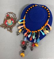 Photo showing two necklaces: a large copper pendant necklace with colorful enamel and an artistic figure, next to a multi-colored bead necklace with dangling elements.