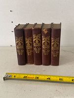 Five miniature leather-bound books displaying ornate gold embossed covers and spines, arranged with a measuring tape showing approximately 3 x 4 inches each.