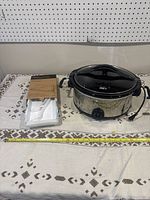 Wide view showing the stainless steel slow cooker with black lid and handles, next to white coating trays and universal lid packaging, all placed on a patterned cloth with measuring tape showing dimensions