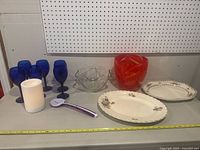 Lot spread showing six cobalt blue stemware glasses, two floral decorative plates, clear glass bowl, red glass item, white candle holder, and purple handled can opener.
