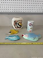 Five decorative fish-themed items placed on a shelf including two vases, two wall plaques shaped like fish, and a fish-shaped bowl or dish, all with paint wear and signs of age.