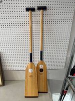 Two wooden paddles displayed standing upright against pegboard, showing full length and overall condition.