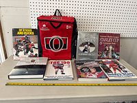 Photo showing six hockey books and one red Ottawa Senators cooler bag with Molson Canadian branding, arranged on a flat surface with a tape measure for scale.