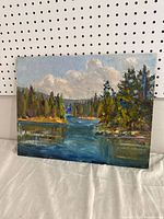 Full view of the oil painting showing a lake scene surrounded by trees and a cloudy sky, displayed against a pegboard.