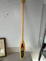 Full view of the wooden paddle standing upright against a pegboard wall, displaying the entire length and design.