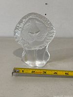 Front view of clear crystal lion paperweight showing detailed facial features and mane with measuring tape indicating about 4 inches tall.