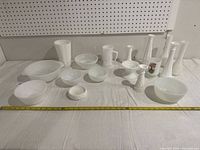 Full view of all 17 milk glass pieces including bowls, pitchers and candlesticks arranged on white cloth with measuring tape showing size of largest bowl.