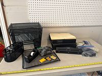 Photo showing a collection of black mesh desk organizers: letter tray, file organizer, pencil cup, tape dispenser, stapler, hole punch, and several small office supplies spread out on a table with measuring tape.