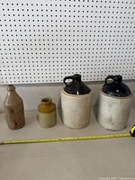 Four old crocks and jugs of varying sizes and colors, lined up with measuring tape showing approximate sizes.